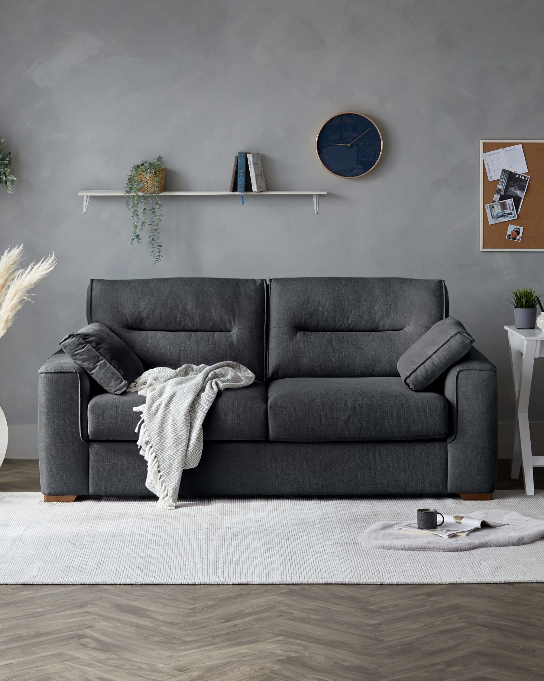 A contemporary dark grey fabric sofa with a plush back, cushioned arms, and matching throw pillows, accented with a light grey knit throw blanket, staged on a white textured area rug. The minimalist styling includes a white floating shelf adorned with books, greenery, and a circular wall clock, beside a corkboard with notes and a white round side table with minimal décor.