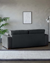 A contemporary charcoal grey fabric sofa with a chaise lounge on the left side, featuring plush back cushions and a clean-lined silhouette. The couch sits on short, round, wooden legs and is presented on a neutral-toned area rug with dark borders.