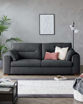 A modern, dark grey upholstered three-seater sofa with plump cushions and armrests, adorned with a mix of decorative pillows in blush, cream, and patterned black and white. In front of the sofa is a small glass-top coffee table with black metal frame, holding books. The room also features a light grey area rug, partially extending under the sofa.