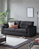 Contemporary charcoal-grey upholstered three-seater sofa with a variety of textured throw pillows in shades of dusty pink, black and white, and off-white. The sofa is accompanied by a modern dark wood and black metal rectangular coffee table displaying books and a ceramic cup. Beneath both, a large area rug with a geometric grey and off-white design anchors the space. A minimalist floor lamp with a unique spherical design stands to the right, adding a touch of elegance.