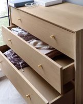 Modern wooden chest of drawers with a light oak finish and sleek, golden handles. The top drawer is partially closed, exposing neatly folded clothing items within, while the second drawer is fully open. Textured wood grain is visible throughout, complementing the minimalist aesthetic. A book and a pair of glasses rest on the smooth top surface, hinting at functionality and lived-in comfort.