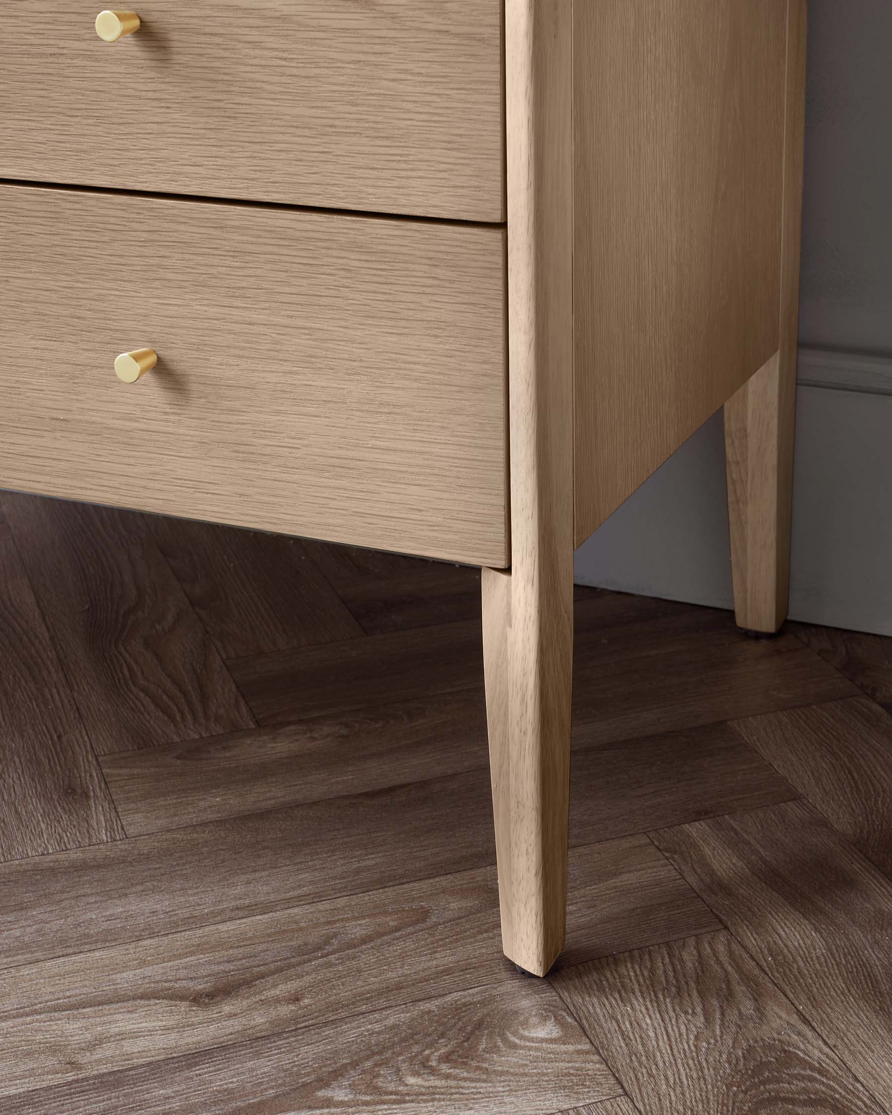 A modern light wood chest of drawers with two drawers featuring round, light yellow knobs, standing on four splayed legs with a clear wood grain finish on a dark wooden floor.