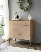 A contemporary light wood dresser with four drawers, featuring round, golden knobs and angular legs, set against a grey wall beside a window with white drapes. A wreath hangs above, and the dresser is adorned with a black vase, a white sculptural piece, and two stacked books.