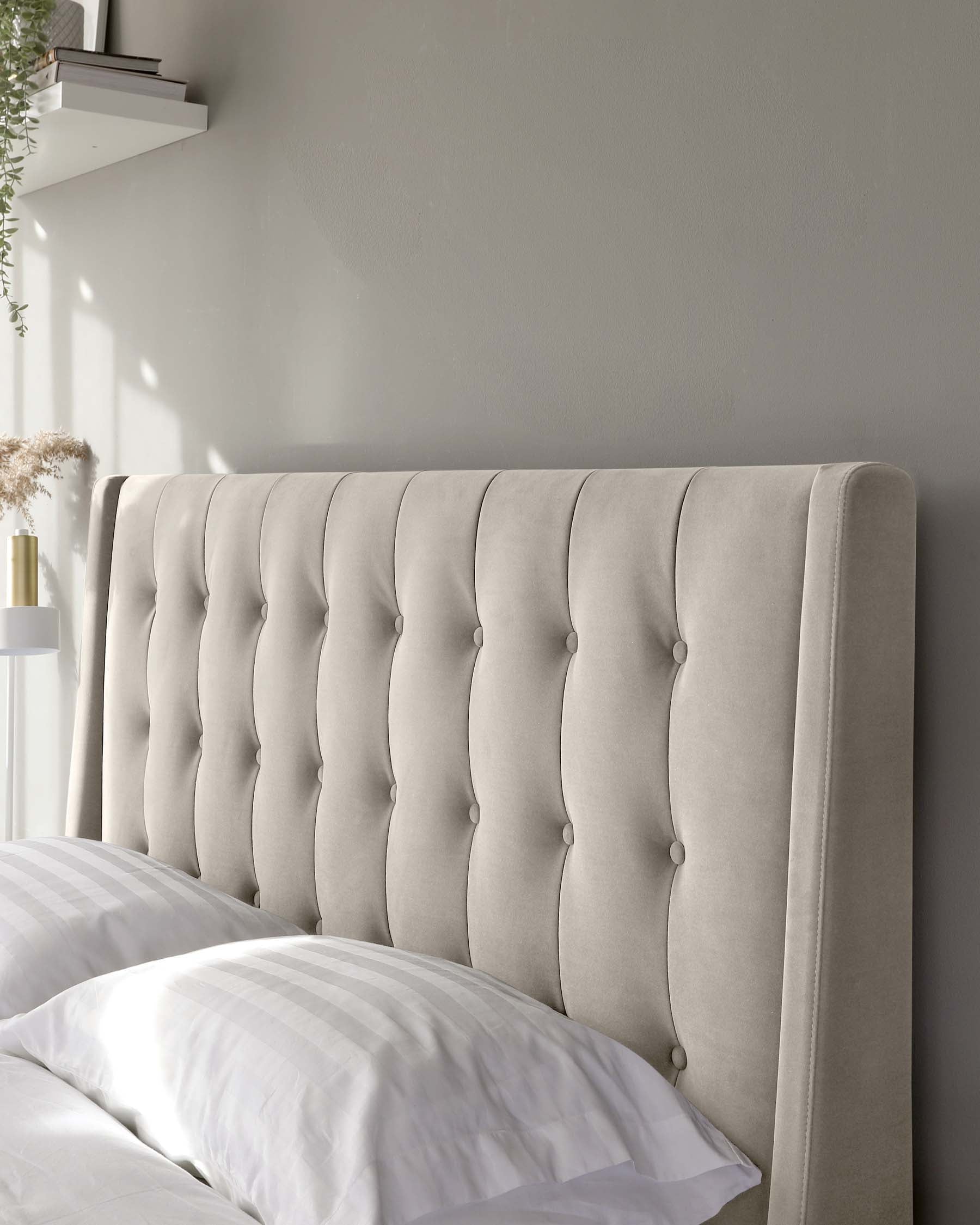 Elegant tufted upholstered headboard in a neutral beige fabric with a vertical channel design and button detailing, mounted against a light grey wall, partially obscured by a white bedding set.