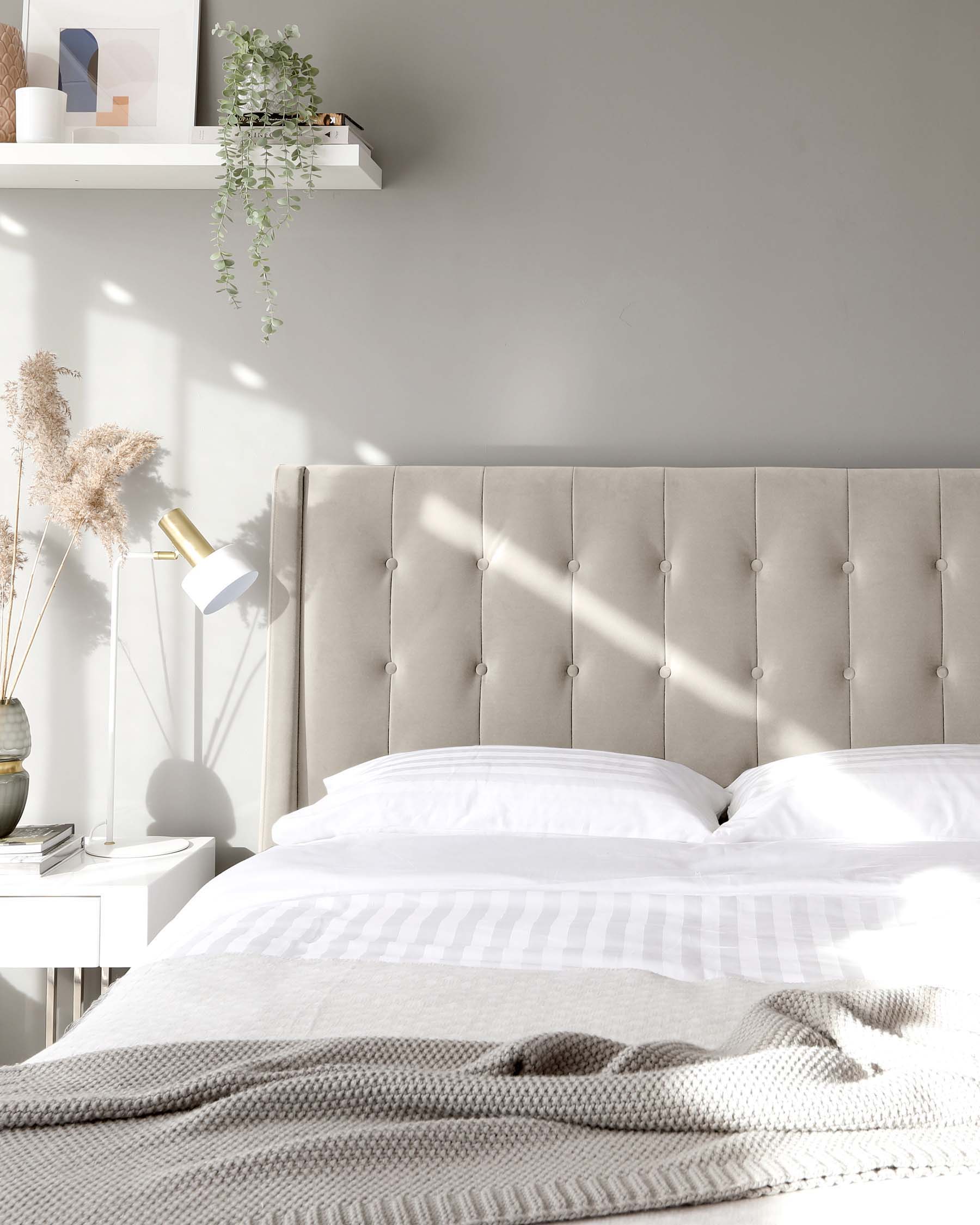 Elegant bedroom featuring a tufted upholstered headboard in a neutral tone, crisp white bedding with subtle striping, and a cosy grey knit throw blanket. A sleek white bedside table supports a modern white lamp, with natural decorative elements and a white floating shelf overhead housing minimalist decor and greenery.