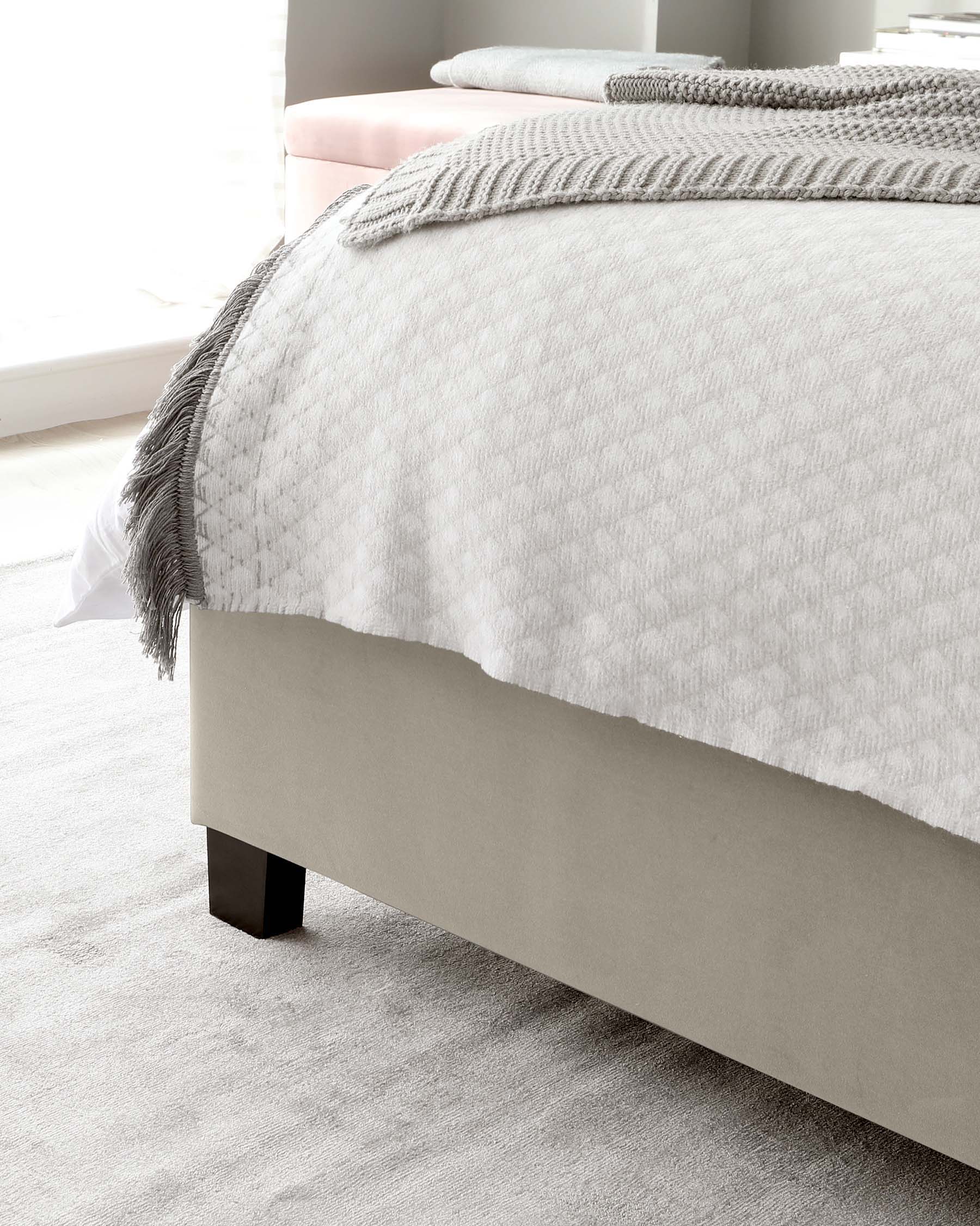 Elegant contemporary bed frame with a minimalist design, upholstered in light grey fabric, featuring clean lines and dark square legs. The bed is adorned with a plush white comforter and a chunky knit grey throw.