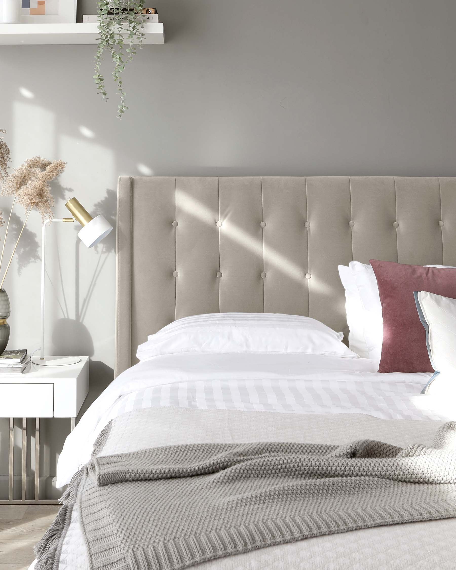 Contemporary bedroom featuring a large upholstered headboard with button tufting in a neutral tone, flanked by a minimalist white nightstand. The bed is adorned with crisp white bedding and a textured grey throw, alongside a burgundy accent pillow, creating an inviting, modern aesthetic.