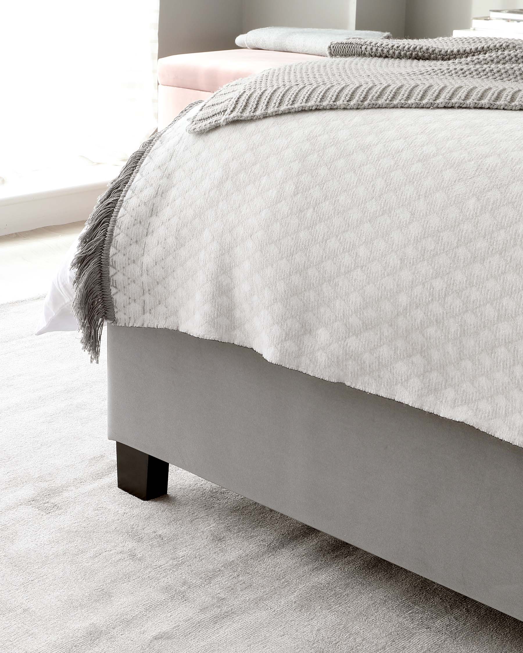 Contemporary-style bed with upholstered headboard and bed frame in light grey fabric, accompanied by a tidy, textured white bedding set with a knitted grey throw on top. The bed sits on a subtle, plush beige carpet.