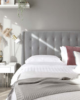 Elegant modern bedroom featuring a large grey upholstered headboard with button-tufted details, accompanied by a sleek white nightstand and a reflective chrome-finished side table. A cosy white and grey striped bedding is topped with a textured grey throw blanket to complete the sophisticated look.