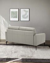 A modern minimalist light grey three-seater sofa with a sleek silhouette, featuring clean lines and cushioned backrests. The sofa sits on slender black tapered legs, positioned on a contemporary two-tone grey area rug.