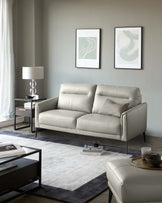 Modern living room setup featuring a sleek light grey upholstered three-seater sofa with cushioned armrests and back, flanked by a dark-framed side table with an elegant table lamp. In front of the sofa is a contemporary rectangular coffee table with a black metal frame and clear glass top on a two-tone area rug. A coordinating ottoman with a similar grey upholstery is placed to the side.