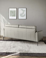 A contemporary light grey leather sofa with a tufted backrest and sleek metal legs, set against a grey wall with two framed abstract art prints, resting on a gradient grey area rug.