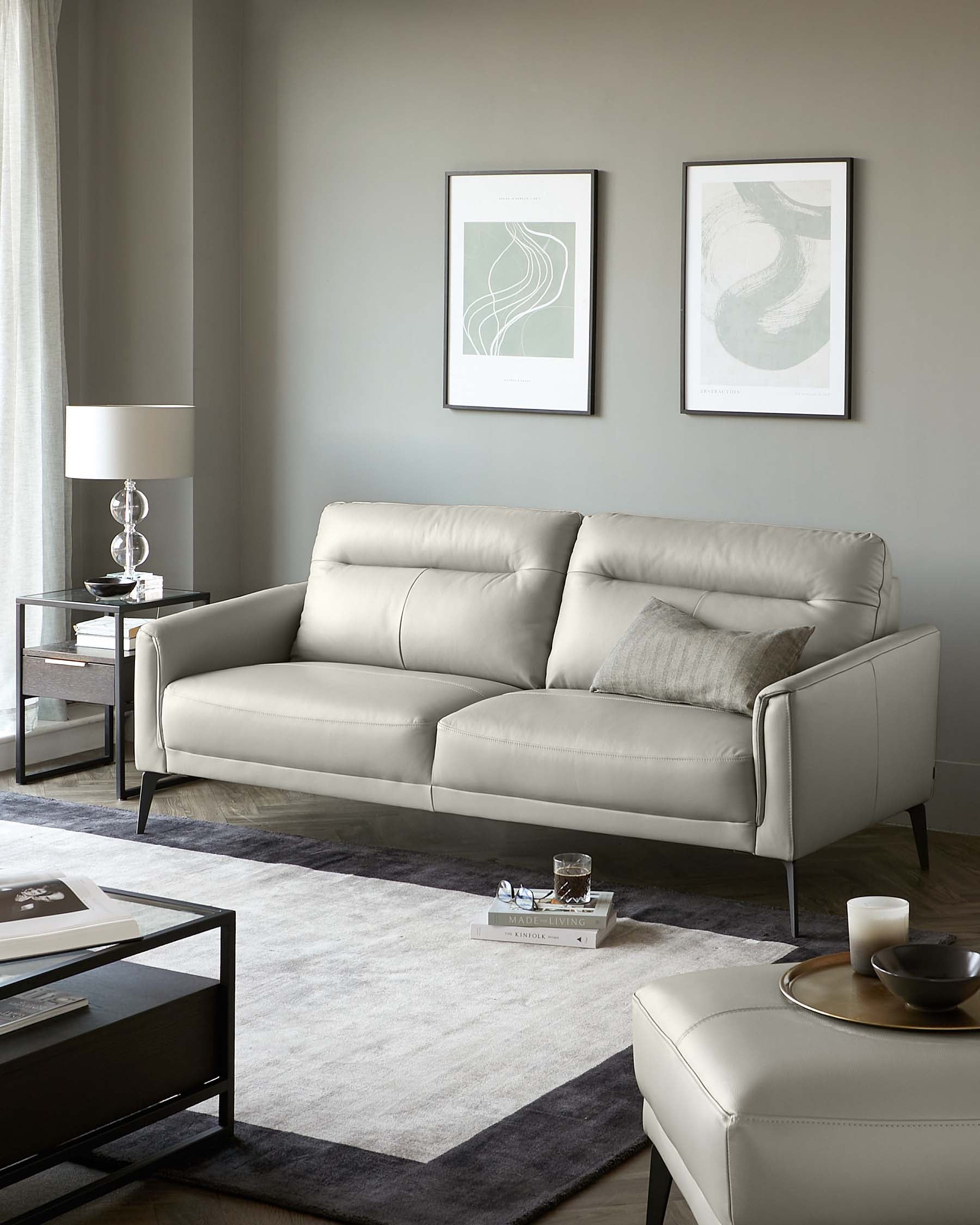 Modern living room set featuring a sleek light grey leather sofa with clean lines and tailored cushions, accompanied by a square black metal and glass side table with a matching coffee table, both displaying simple, straight lines. Decorative elements include a neutral-toned area rug, a cylindrical white lamp with a round shade, and two framed abstract wall art pieces adding visual interest to the space.