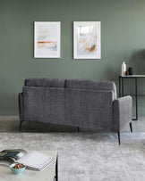 Contemporary grey fabric sofa with minimalist design and dark wooden legs, accompanied by a sleek, black, rectangular side table with a minimalist vase and bottle on top, set against a muted green wall with abstract art.