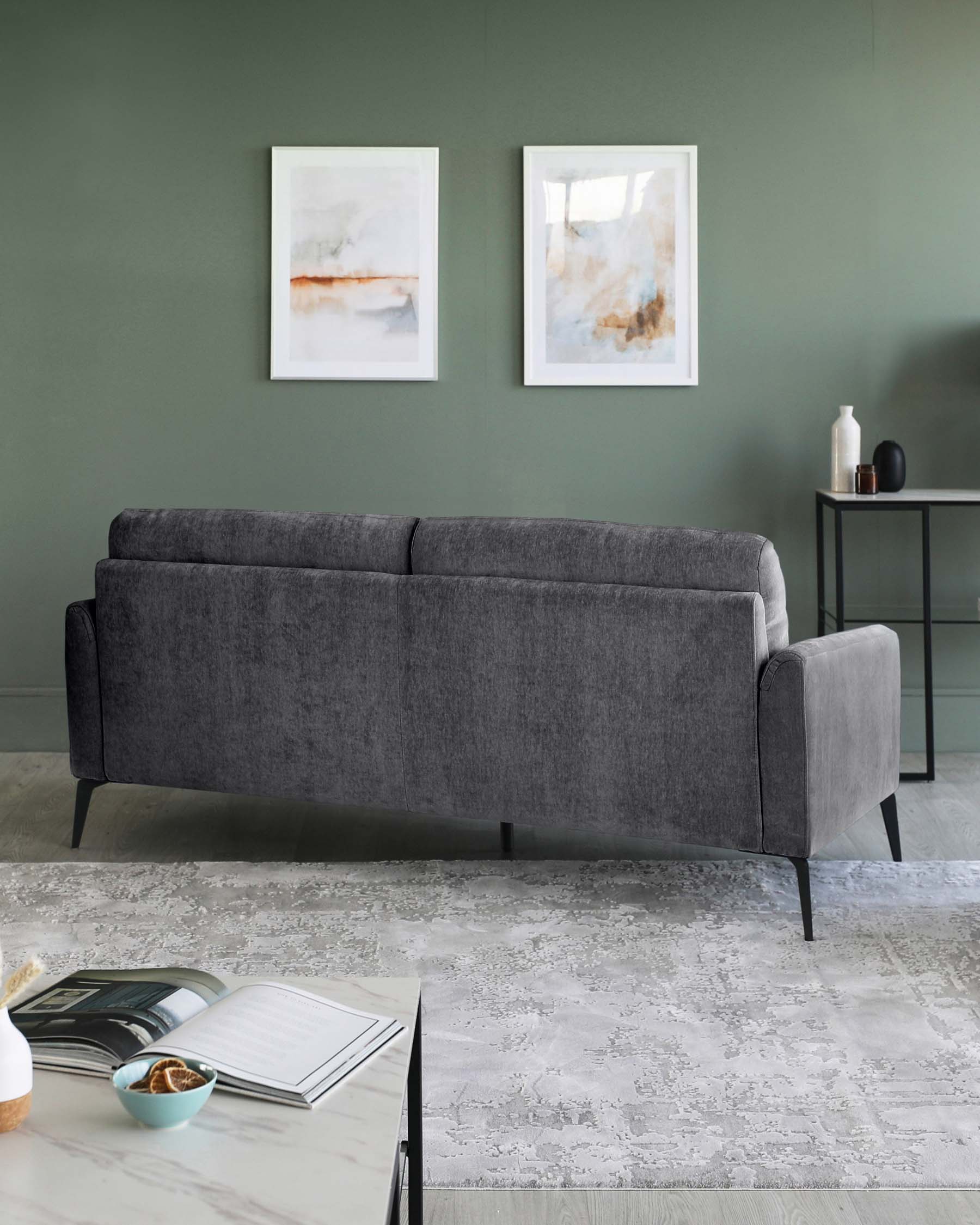 Contemporary dark grey upholstered sofa with a streamlined design and rounded corners sitting on black tapered legs, accompanied by a minimalist circular black side table with a simple vase and a bowl atop. The furniture is presented on a textured white and grey area rug, against a muted green wall adorned with two abstract framed art pieces.