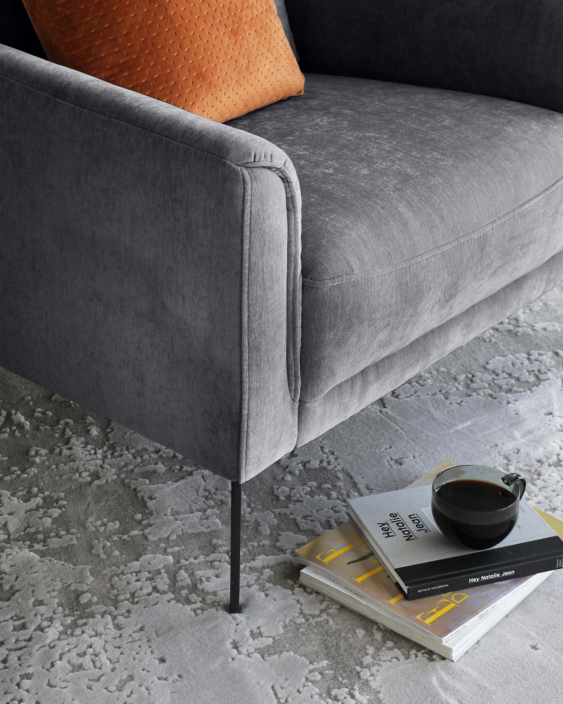 Contemporary grey upholstered sofa with a plush seat cushion, sleek armrest, and black metal legs, accompanied by an orange textured throw pillow. A stack of books and a black coffee cup rest on a grey patterned area rug beside the sofa.