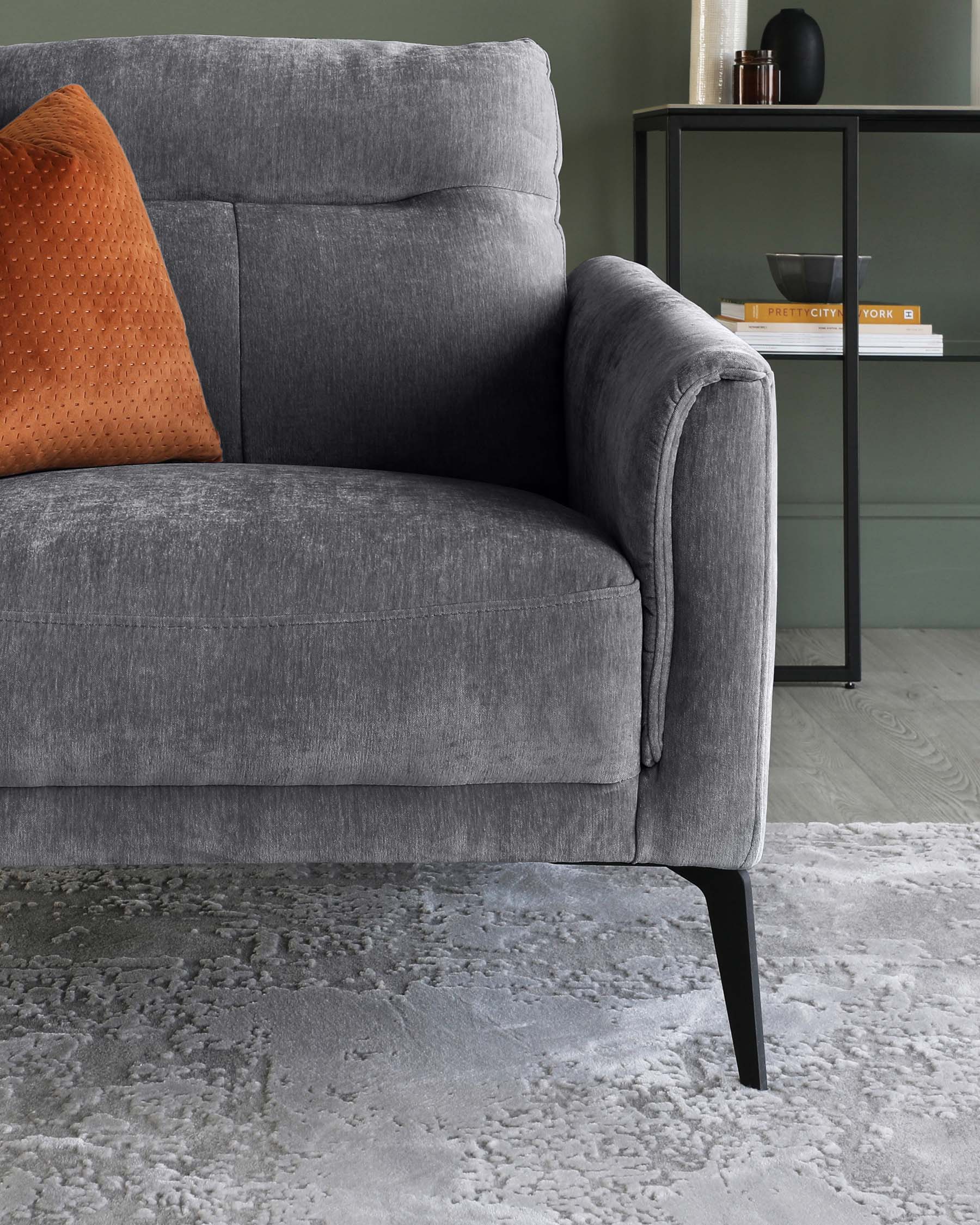 A modern charcoal grey fabric sofa with a plush back cushion and an orange textured throw pillow, standing on sleek black tapered legs, beside a minimalist black metal side table with books and decorative items. The scene is completed by a patterned grey area rug.