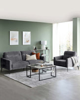 Elegant living room furniture set against a muted green wall, featuring a dark grey fabric sofa and matching armchair with sleek black metal legs. A rectangular glass-top coffee table with a black metal frame and a lower wooden shelf sits on a textured grey area rug. A side table with similar design elements and a round top is positioned by the sofa, complemented by a modern table lamp with a white shade.