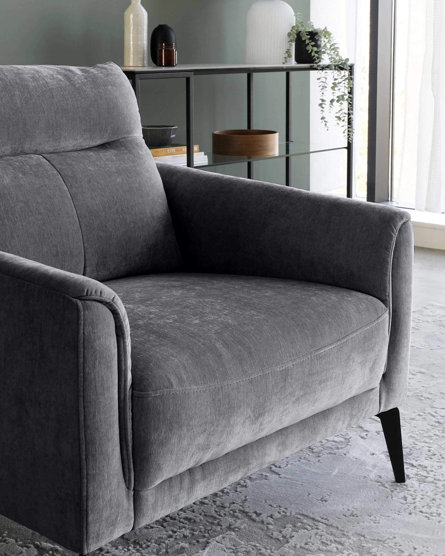 Contemporary grey fabric loveseat with a streamlined design, featuring plush back cushion, flared armrests, and black tapered wooden legs. A sleek glass-fronted shelving unit is visible in the background displaying decorative vases, books, and potted plants.