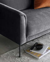 A contemporary dark grey upholstered sofa with clean lines and thin metal legs, paired with a small white rectangular coffee table on which rests a black ceramic cup and saucer and a stack of hardcover books. The setting is complemented by a textured grey area rug underneath.