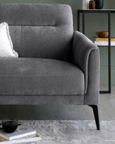 Elegant grey fabric sofa with clean lines and a contemporary design. Features plush cushioning, a single white decorative pillow, and sleek black metal legs. Positioned next to a modern, minimal glass shelving unit displaying books and decorative items. Set against a muted green wall, over a textured grey area rug, creating a sophisticated and cosy atmosphere.