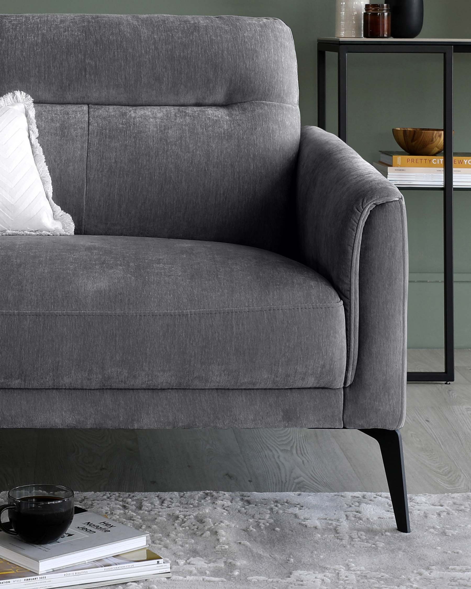 Elegant dark grey upholstered loveseat with structured armrests and a tailored silhouette on slender black metal legs, accented with a single decorative white textured throw pillow. A minimalist black metal and glass display shelf stands in the background with decorative items, complementing the modern aesthetic.