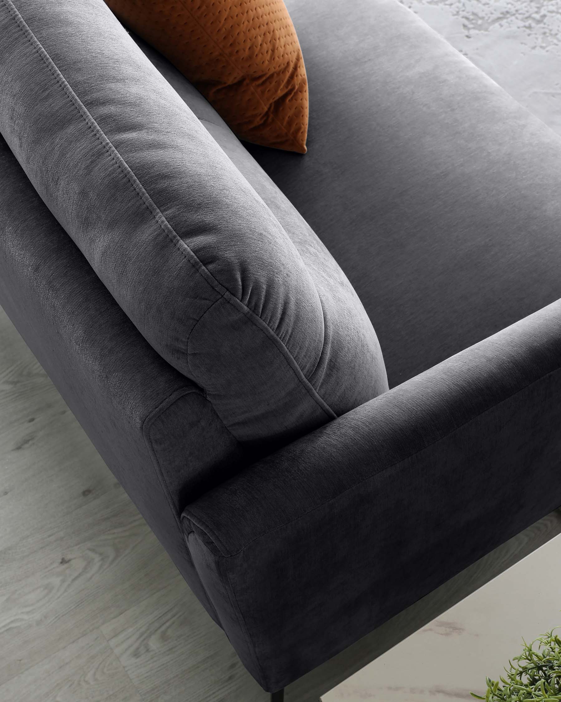 Close-up of a modern charcoal grey sofa with a plush texture and a single burnt orange decorative pillow. The sofa features clean lines, a thick padded seat cushion, a rounded armrest, and an elevated backrest with a cylindrical bolster cushion. It appears to be set on a subtle angled leg design, with a light wood floor and a hint of a green plant in the foreground.