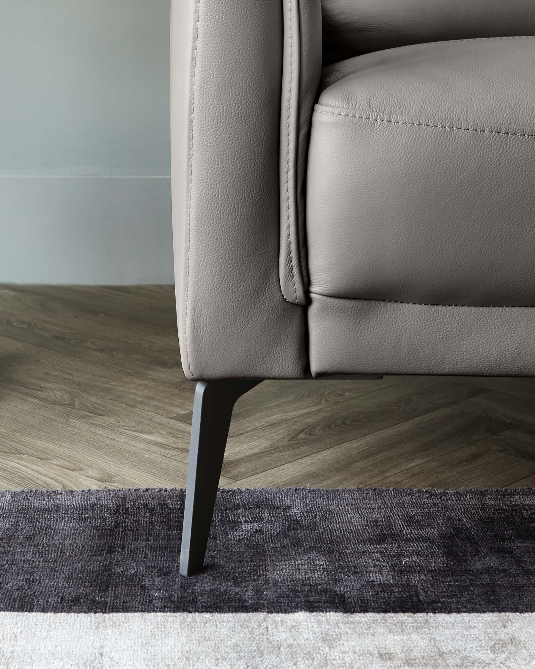 Close-up of a modern grey sofa with a textured upholstery and a sleek, black, tapered leg design, standing on a two-tone area rug with grey and deep purple hues over a wooden floor.