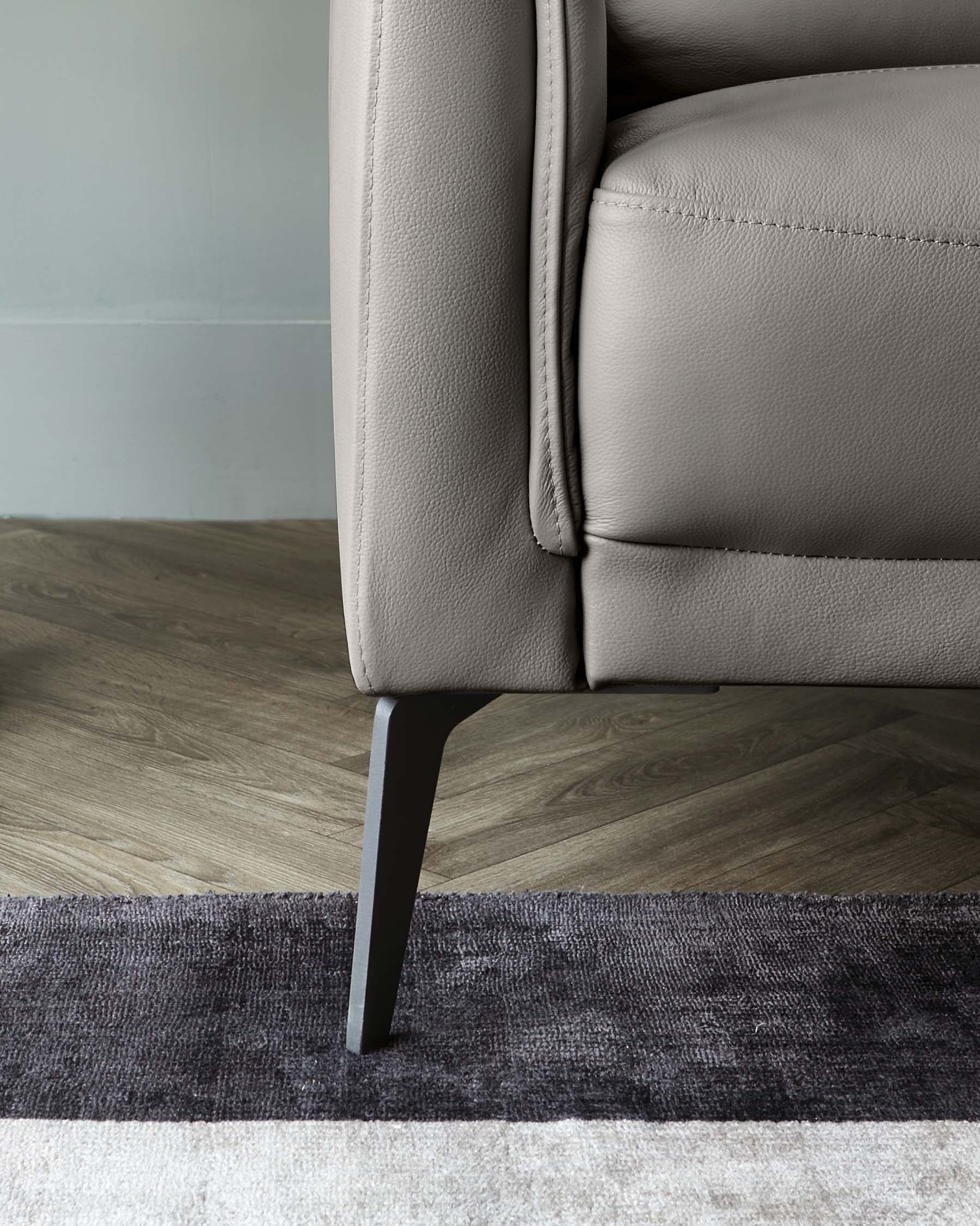 A modern taupe leather sofa with clean lines and a single visible, slender, black angled leg resting on a two-tone grey area rug, set against a hardwood floor and a light grey wall.