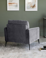 Modern charcoal grey fabric armchair with a sleek, boxy silhouette and black angular wooden legs, positioned on a muted grey and white patterned area rug.