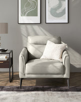 Elegant modern light grey upholstered armchair with a plush seat cushion and a decorative white textured throw pillow, featuring sleek black metal legs. Beside it is a contemporary style square side table with a black frame and a dark wooden top, accessorized with a small silver bowl. The decor is complemented by two abstract wall art pieces and a soft-focus grey area rug on a wooden floor, creating a stylish and sophisticated interior setting.