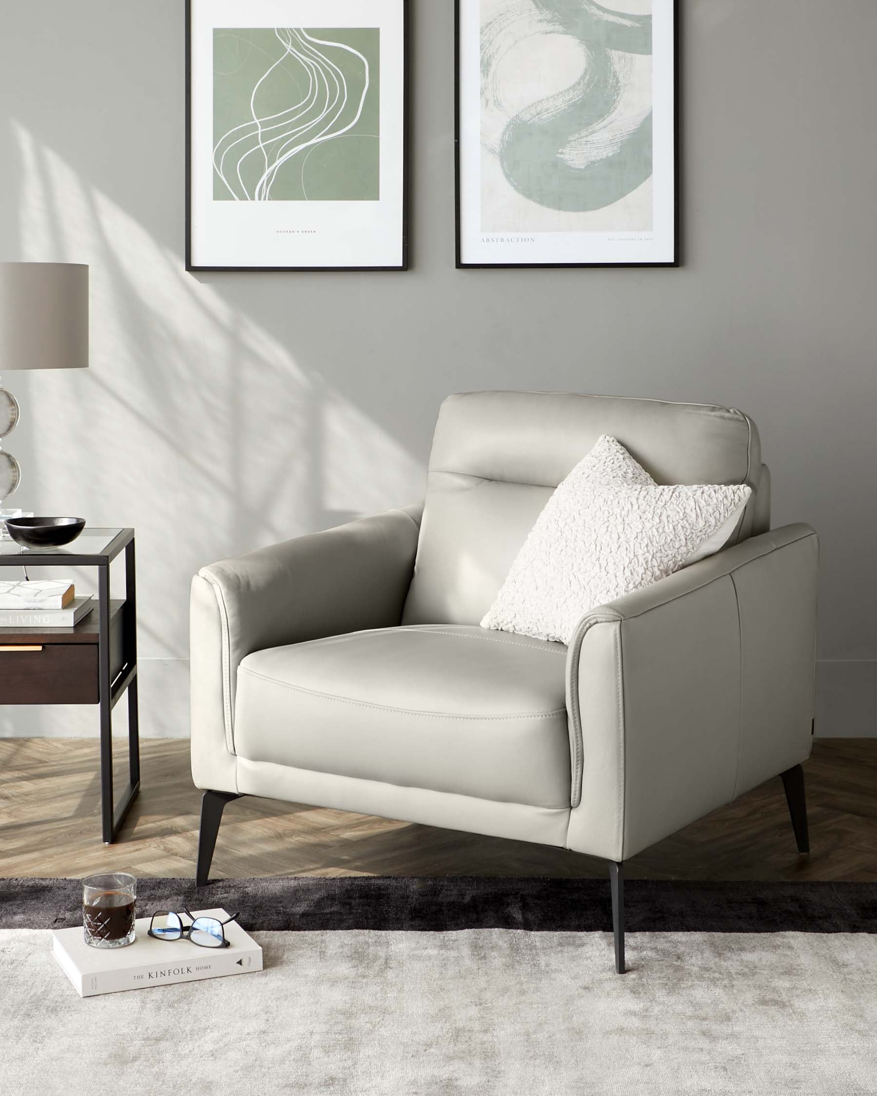 Elegant contemporary style armchair with light grey upholstery and sleek black legs, complemented by a soft white decorative pillow. Adjacent to the chair is a modern black end table with a bottom shelf, featuring decorative items including a lamp, bowl, and books. The setup is arranged on a plush two-tone area rug.