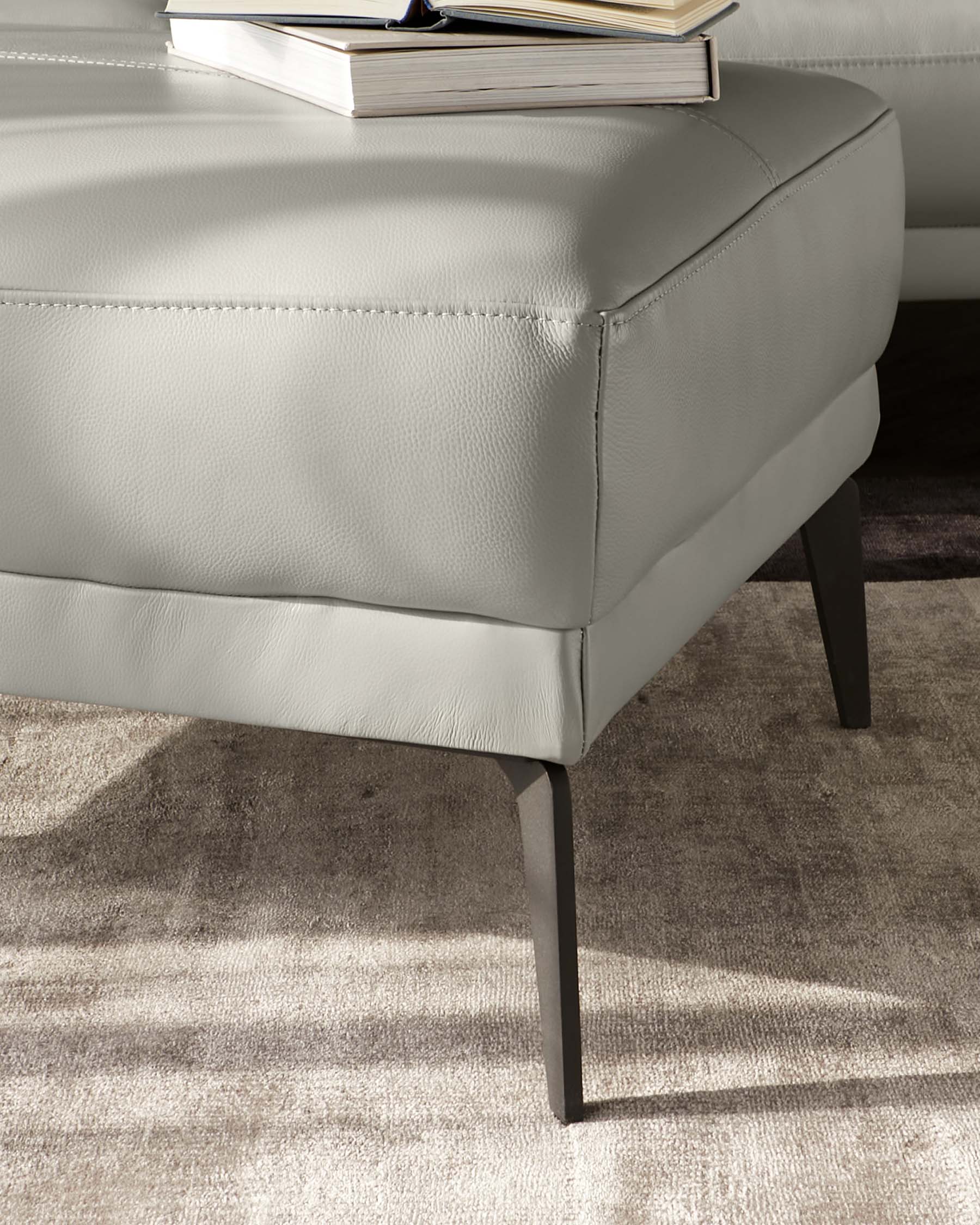 Contemporary grey leather ottoman with clean lines and angular, black tapered legs, partially obscured by a book on top.