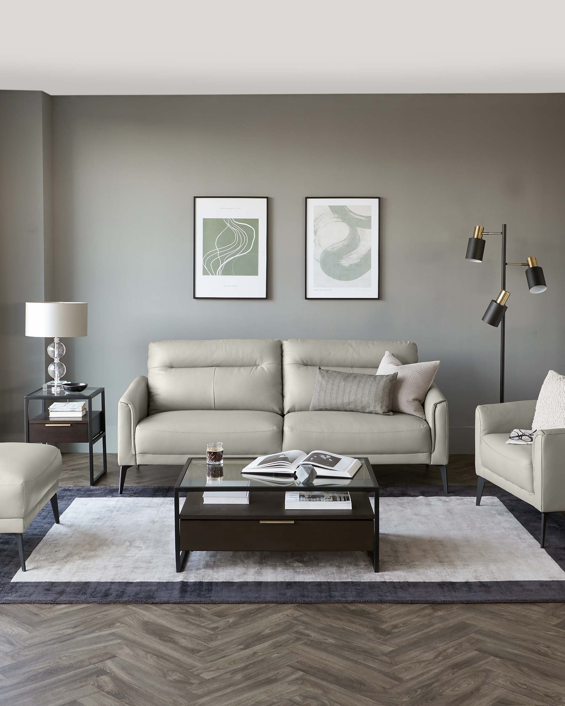 Modern living room set including a light grey upholstered sofa with matching armchair and ottoman, a dark wood rectangular coffee table with a glass top, and a black side table with drawers. Decorative elements include a white table lamp, framed artwork above the sofa, a floor lamp with multiple adjustable arms, and assorted throw pillows. The furniture is arranged on a muted area rug over herringbone-patterned wood flooring.