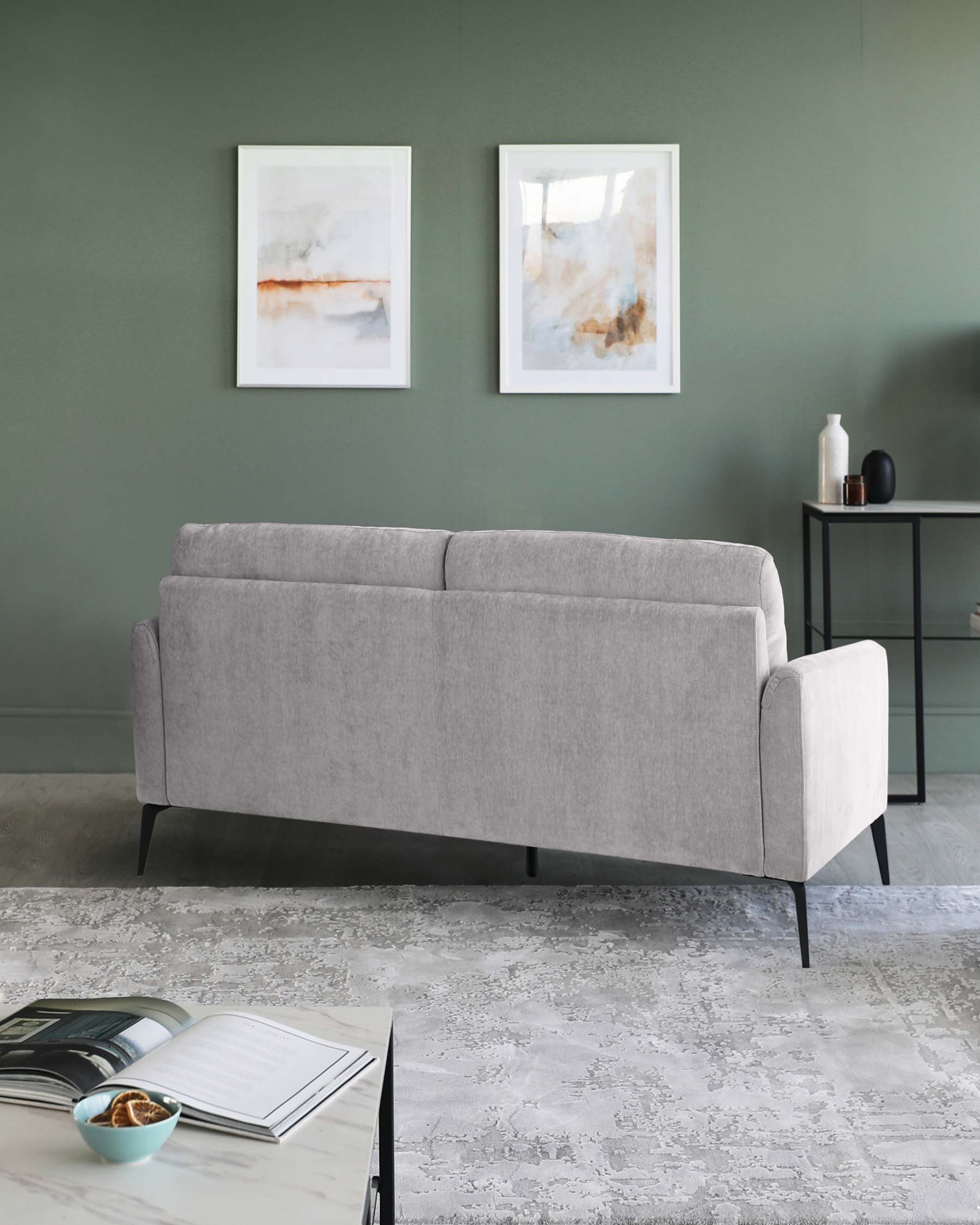 Modern light grey three-seater sofa with sleek black legs, positioned against a dark green wall, complemented by a minimalist black side table with a decorative vase and bottle, and accentuated by two abstract art frames above. The setting is completed with a textured grey area rug and a black coffee table book in the foreground.