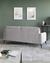 Modern light grey fabric sofa with clean lines and slim, black metal legs, positioned on a textured grey area rug. A minimalist black side table with a round top is placed to the right, accessorized with a white vase and dark decorative pieces.