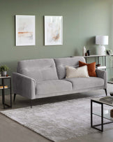 Contemporary three-seater sofa with plush grey upholstery and clean lines, standing on tapered metal legs. Accompanying the sofa is a minimalist black metal end table with a glass top and lower shelf, and a sleek, matching metal console table with a wooden top and two shelves hosting various decorative items and a modern table lamp. Underneath is a large, textured area rug in muted grey and cream tones.