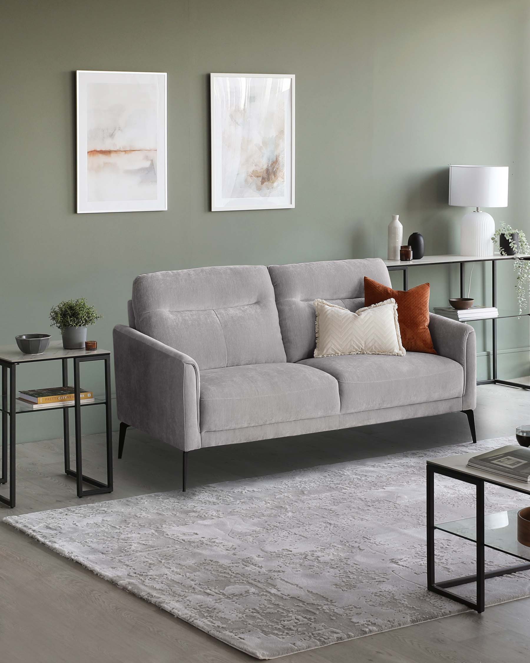 A modern living room featuring a light grey fabric three-seat sofa with clean lines and tapered legs, paired with a textured off-white area rug. Flanking the sofa are two sleek black metal side tables with glass tops, displaying decorative items and books. A minimalist console table with a glass top and metal frame stands against the wall, accented with an assortment of elegant vases and a contemporary white lamp. The space is completed by two abstract framed art pieces on the wall.