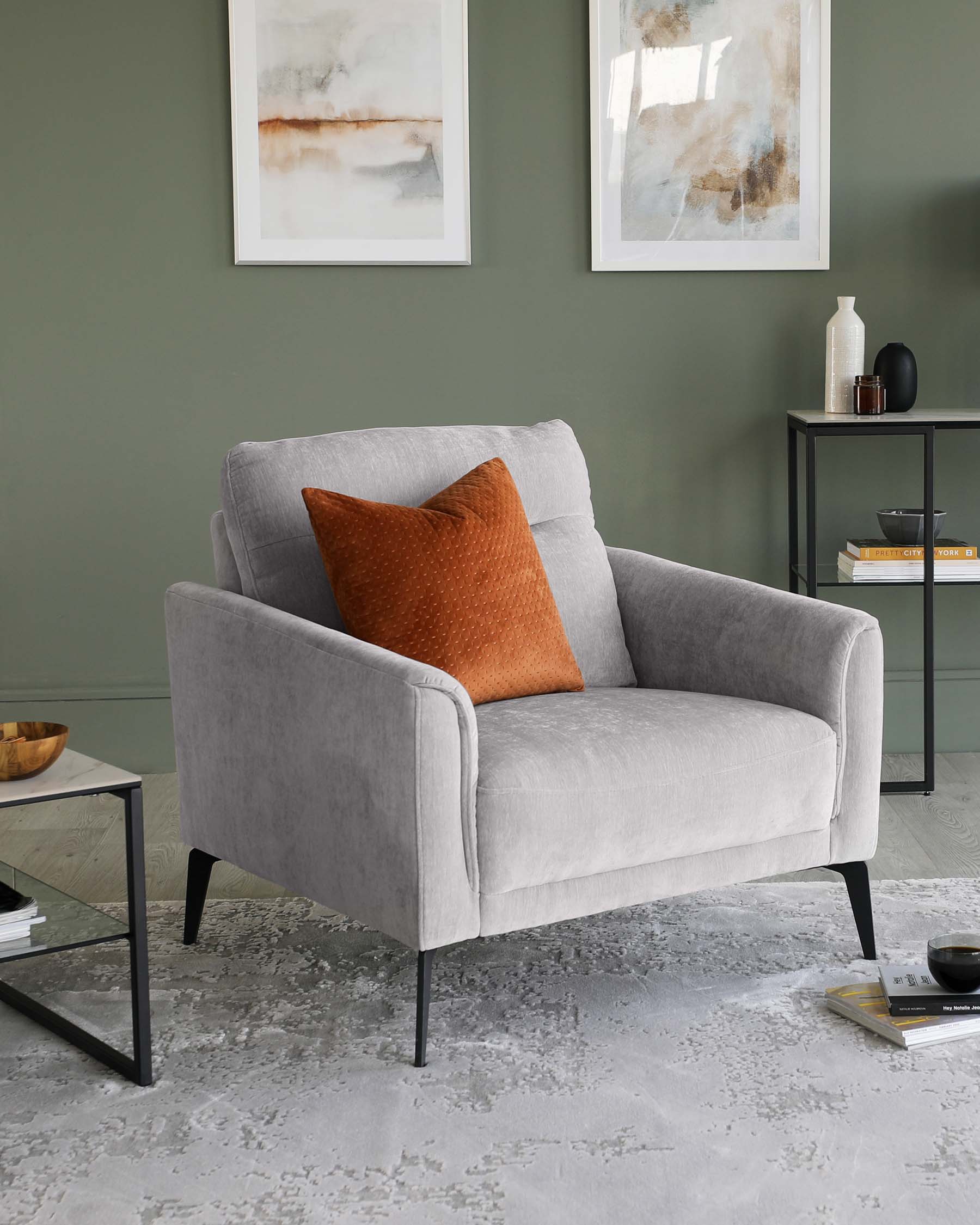 Light grey upholstered armchair with a dark brown leather pillow, featuring clean lines and black metal legs, adjacent to a minimalist black metal side table with a glass top and lower shelf displaying books and decorative items, set against a sage green wall with abstract art and a modern ceramic vase.