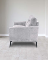 Modern light grey fabric armchair with clean lines and black tapered legs, situated in a minimalist white room with light wooden flooring.