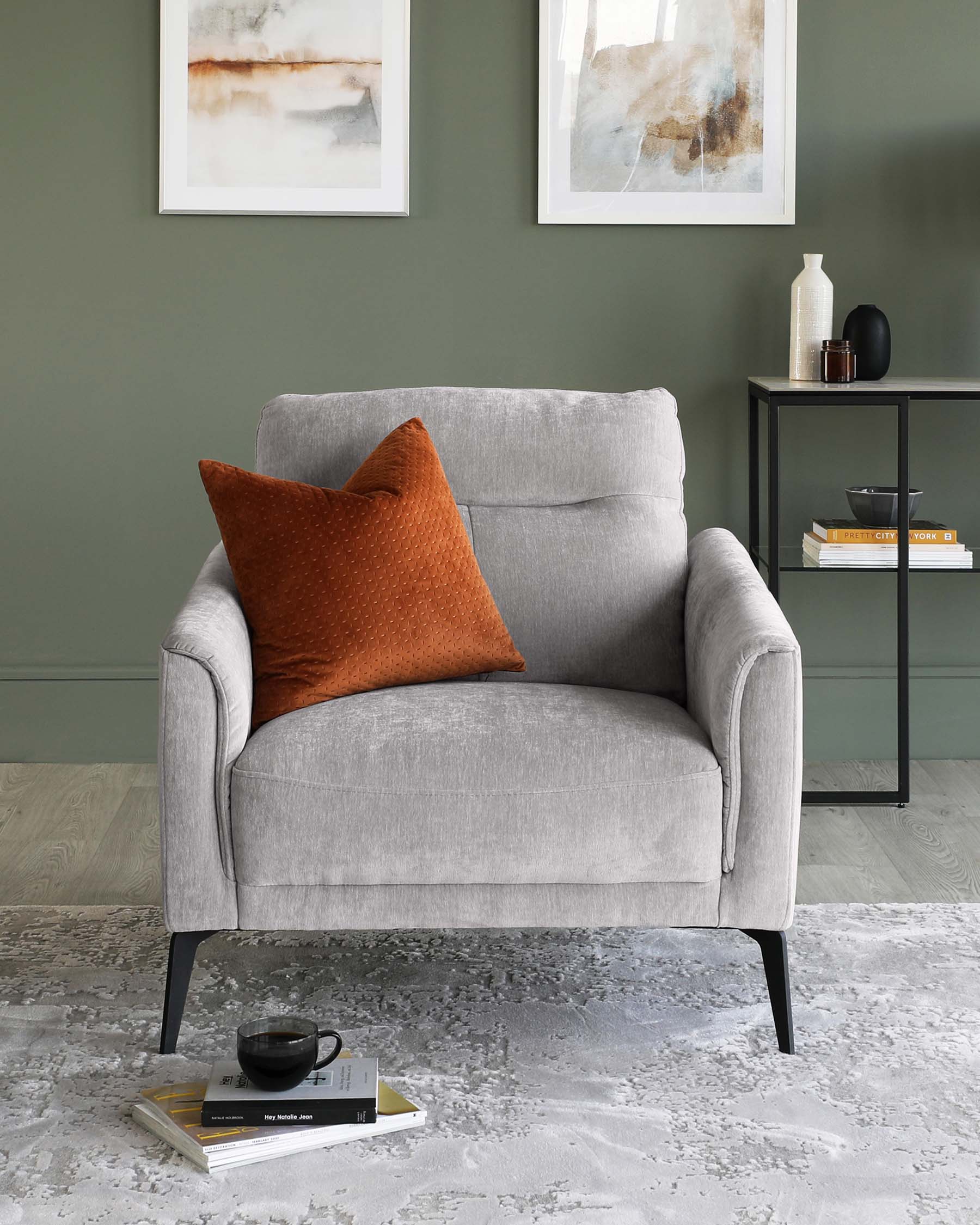 Contemporary light grey fabric armchair with a plush seat, curved armrests, and black metal legs, paired with a brown textured throw pillow. To the side, a modern minimalist black metal side table holds decorative items and books. The setting includes a neutral-toned abstract rug and two framed abstract artworks on a green wall.