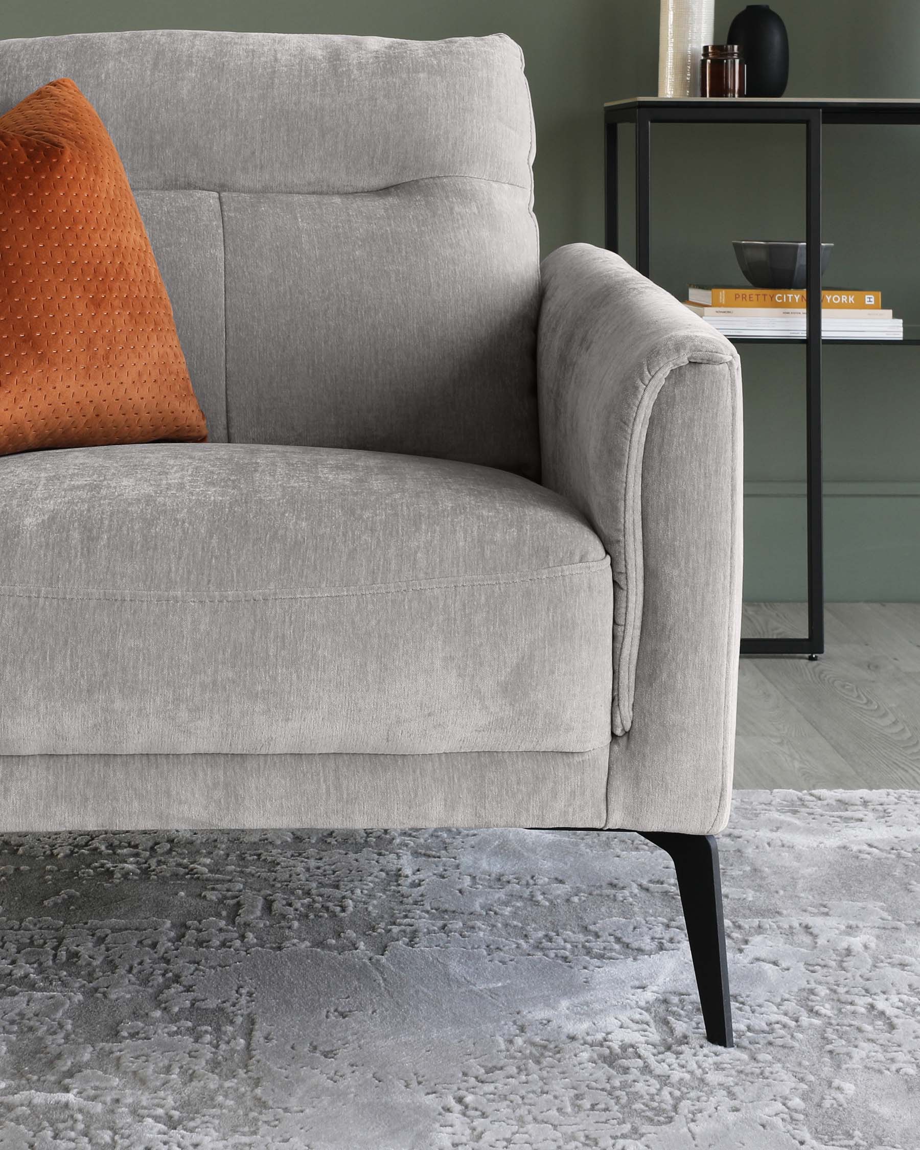 A contemporary grey fabric sofa with a plush back cushion and an orange textured throw pillow. The sofa has a rounded armrest and is supported by tapering black metal legs. Beside it, there's a modern black metal end table with a minimalistic frame, holding decorative items and books. The furniture rests on a textured grey area rug, set against a muted green wall, adding to the modern and cosy ambiance of the space.