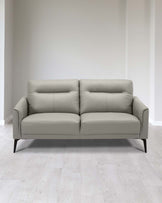 Modern minimalist light grey leather sofa with clean lines, featuring a slightly angled backrest, plush seating, and tapered dark wooden legs. The sofa is set against a neutral backdrop with pale wood flooring.