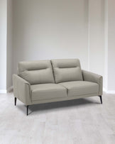 Modern light grey two-seater leather sofa with cushioned backrest and seat, featuring sleek armrests and angled black wooden legs, set in a minimalist room with light wood flooring and white walls.