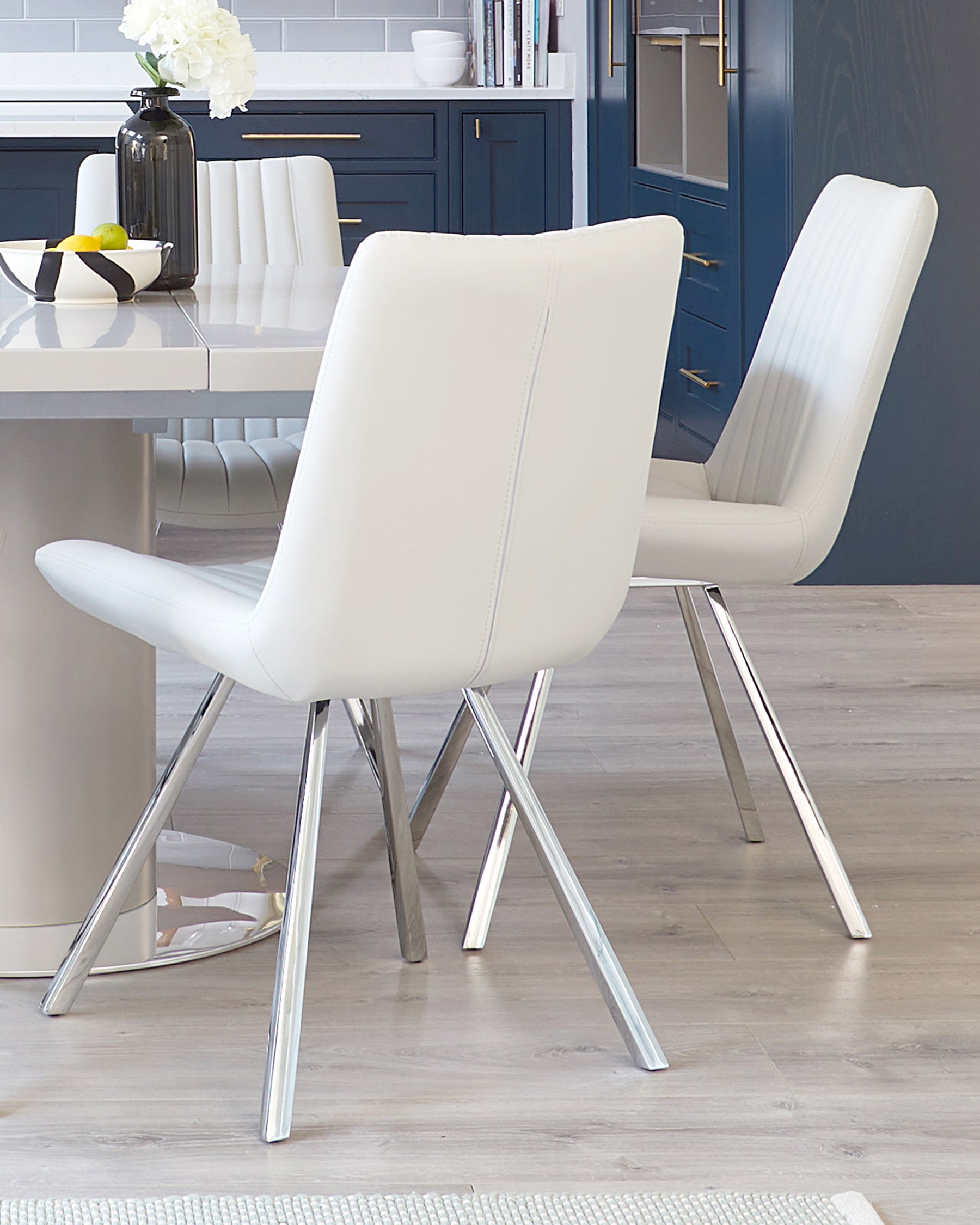 Modern white dining chairs with a sleek design featuring tapered metallic legs and a smooth, curved backrest with a central stitch line. The chairs complement a minimalist white dining table, set against a kitchen with dark blue cabinetry and a hardwood floor.