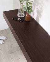 A modern dark wood console table with a rectangular flat top and tapered clear glass side panels, set against a light wall and wood flooring.