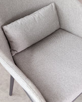 Close-up of a contemporary armchair with a textured light grey fabric upholstery and a rectangular lumbar pillow. The chair features clean lines, an angled backrest, and dark grey metal legs.