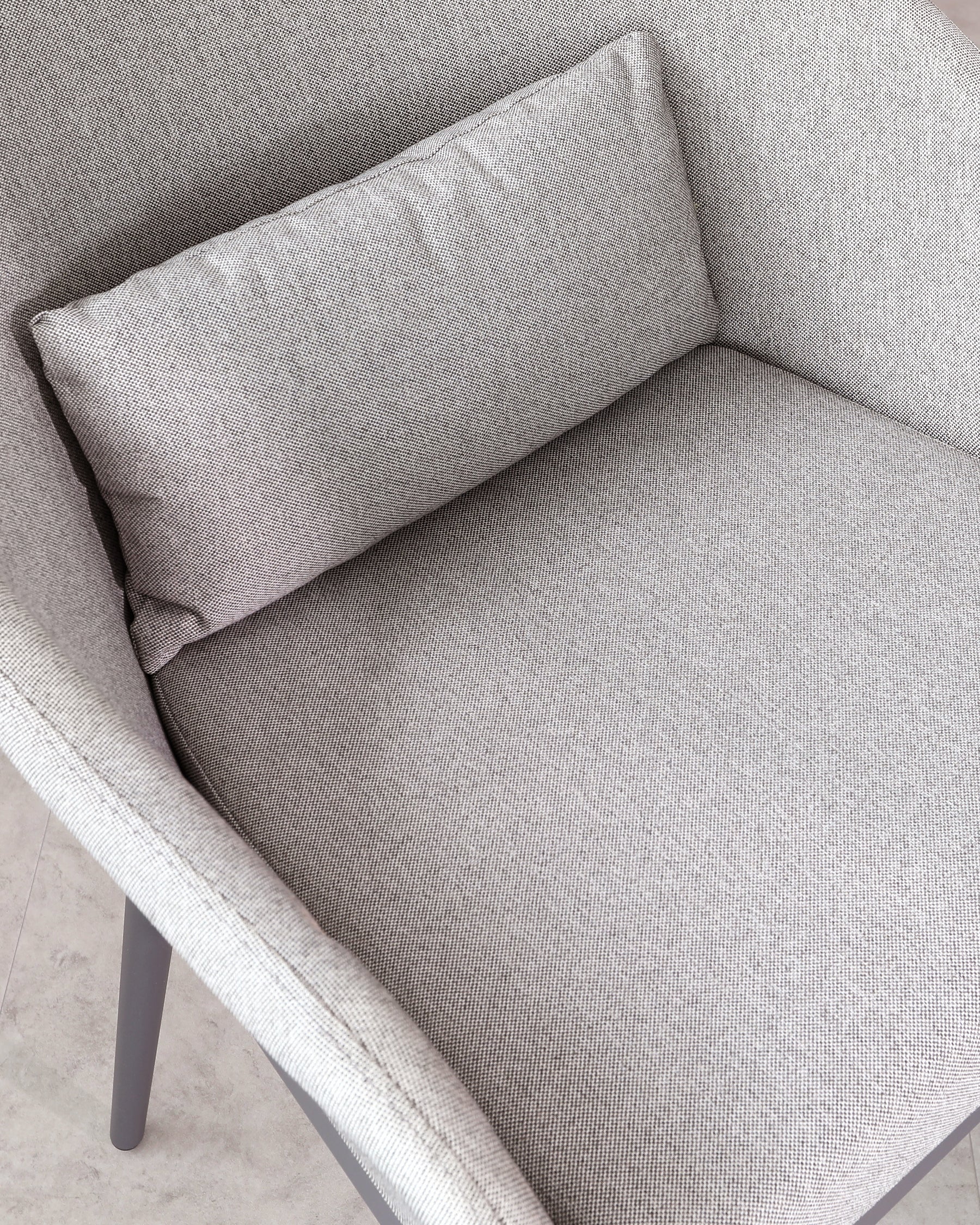 Close-up of a contemporary armchair with a textured light grey fabric upholstery and a rectangular lumbar pillow. The chair features clean lines, an angled backrest, and dark grey metal legs.