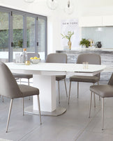 Modern dining room set with a white rectangular table featuring a sleek geometric base and a set of six taupe upholstered chairs with slender metal legs.