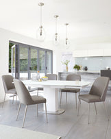 A modern dining room featuring an oval white dining table with a pedestal base, surrounded by six sleek taupe upholstered chairs with slender metal legs.